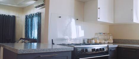 A compact kitchen with a stove, oven, and countertop. There are jars on the counter and a window with curtains.