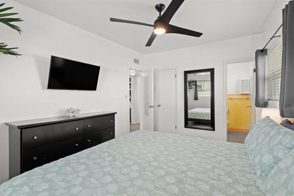 Une chambre à coucher comprenant un lit, un ventilateur de plafond, un téléviseur fixé au mur, une commode et une porte donnant sur une autre pièce.