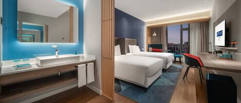 Ein modernes Hotelzimmer mit einem großen Spiegel, einem Waschbecken, zwei Betten, einem Schreibtisch und Blick auf die Stadt.