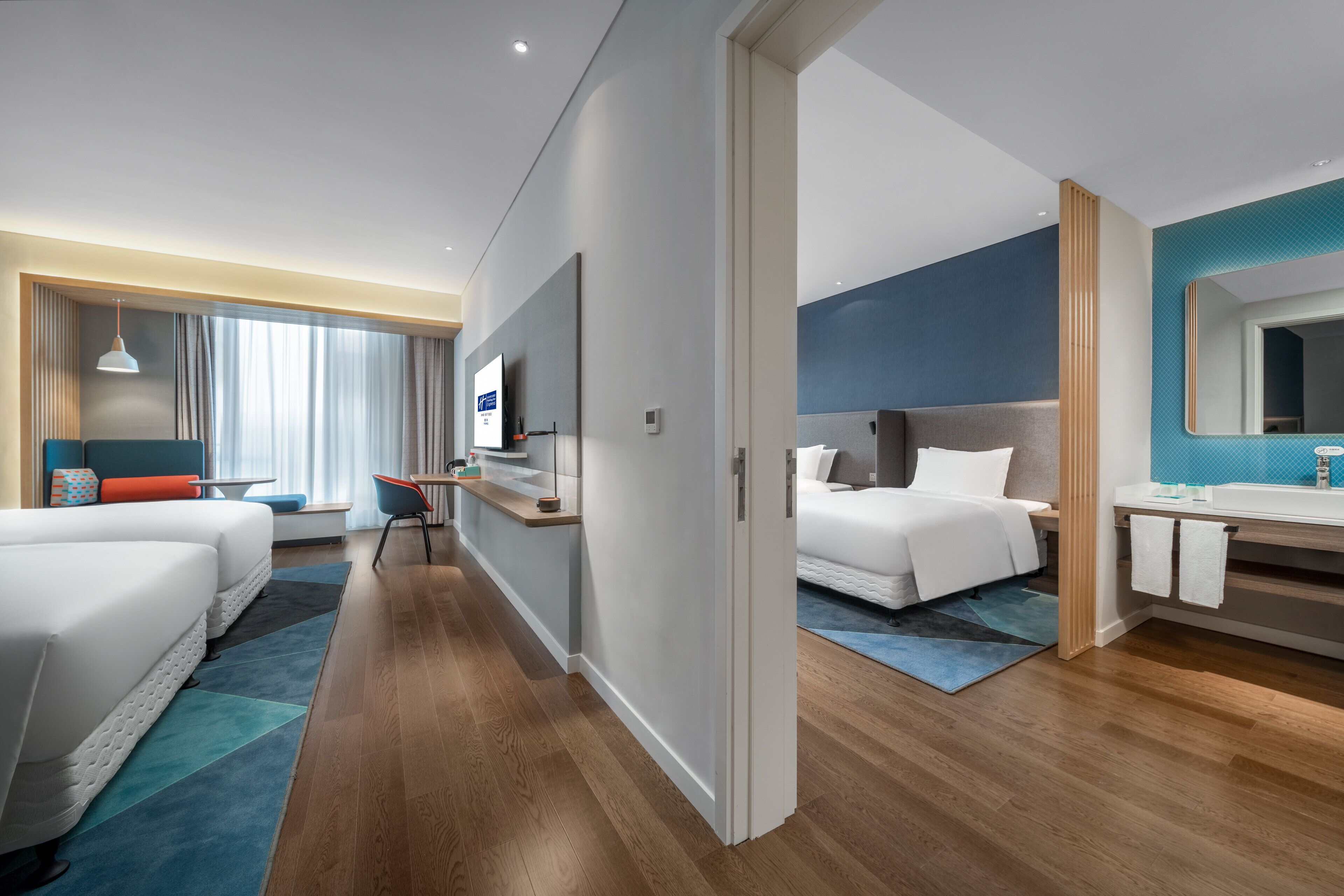 Una habitación de hotel moderna con dos camas, un escritorio, un televisor y un baño con espejo y lavamanos.