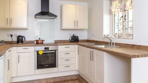 A modern kitchen with white cabinets, a black range hood, a built-in oven, a coffee maker, and a window with floral curtains. - Breckland Cottage: Secluded Country Getaway (Hockwold cum Wilton)