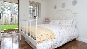 A bedroom with a white bed, a window with blinds, a small bedside table, and a view of a garden. - Breckland Cottage: Secluded Country Getaway (Hockwold cum Wilton)