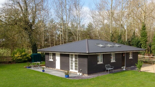A single-story house with a dark exterior, a flat roof, and a small front yard with a lawn and a few potted plants. - Breckland Cottage: Secluded Country Getaway (Hockwold cum Wilton)