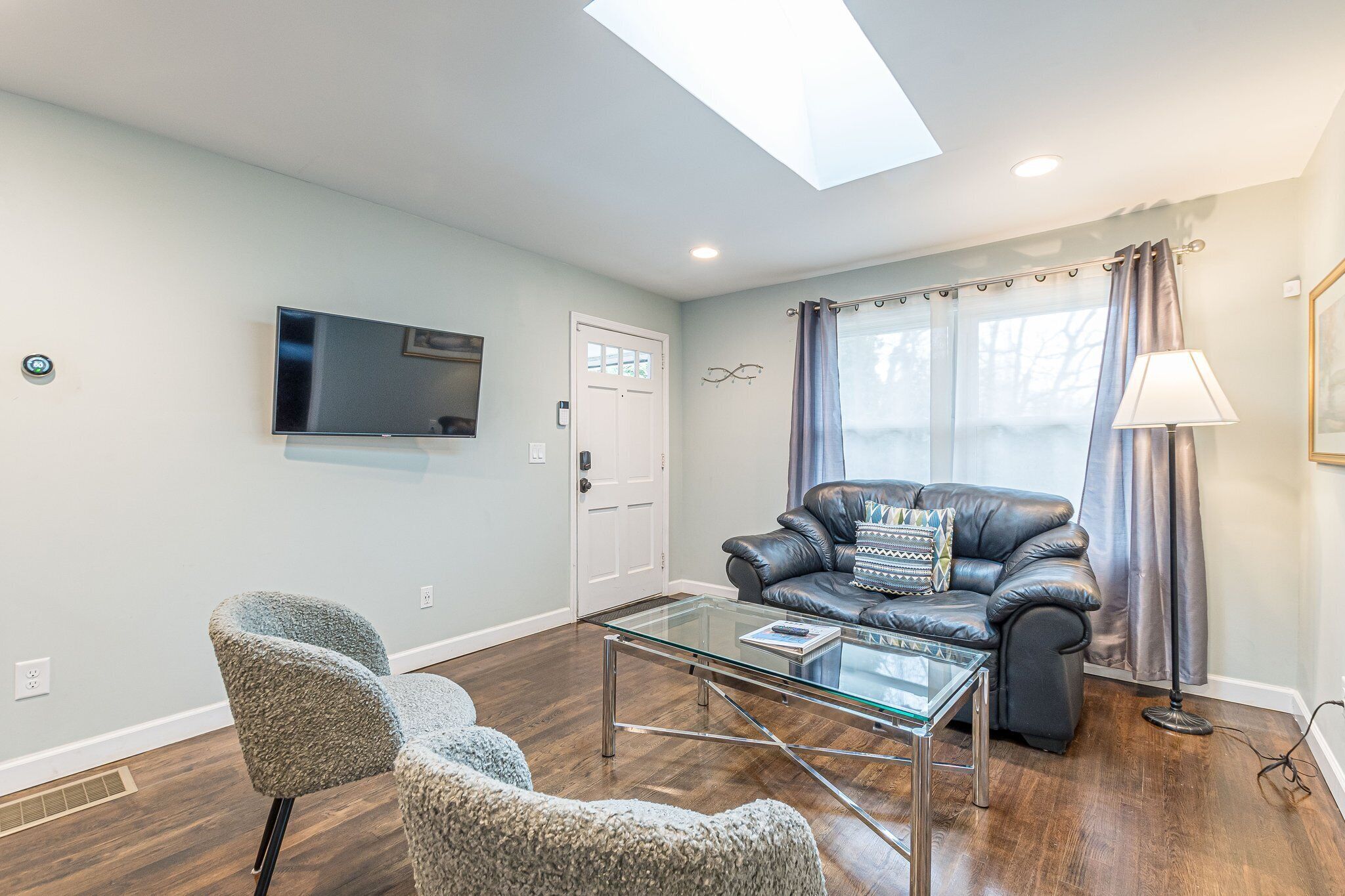 A modern living room with a glass coffee table, leather sofa, and a flat-screen TV mounted on the wall.