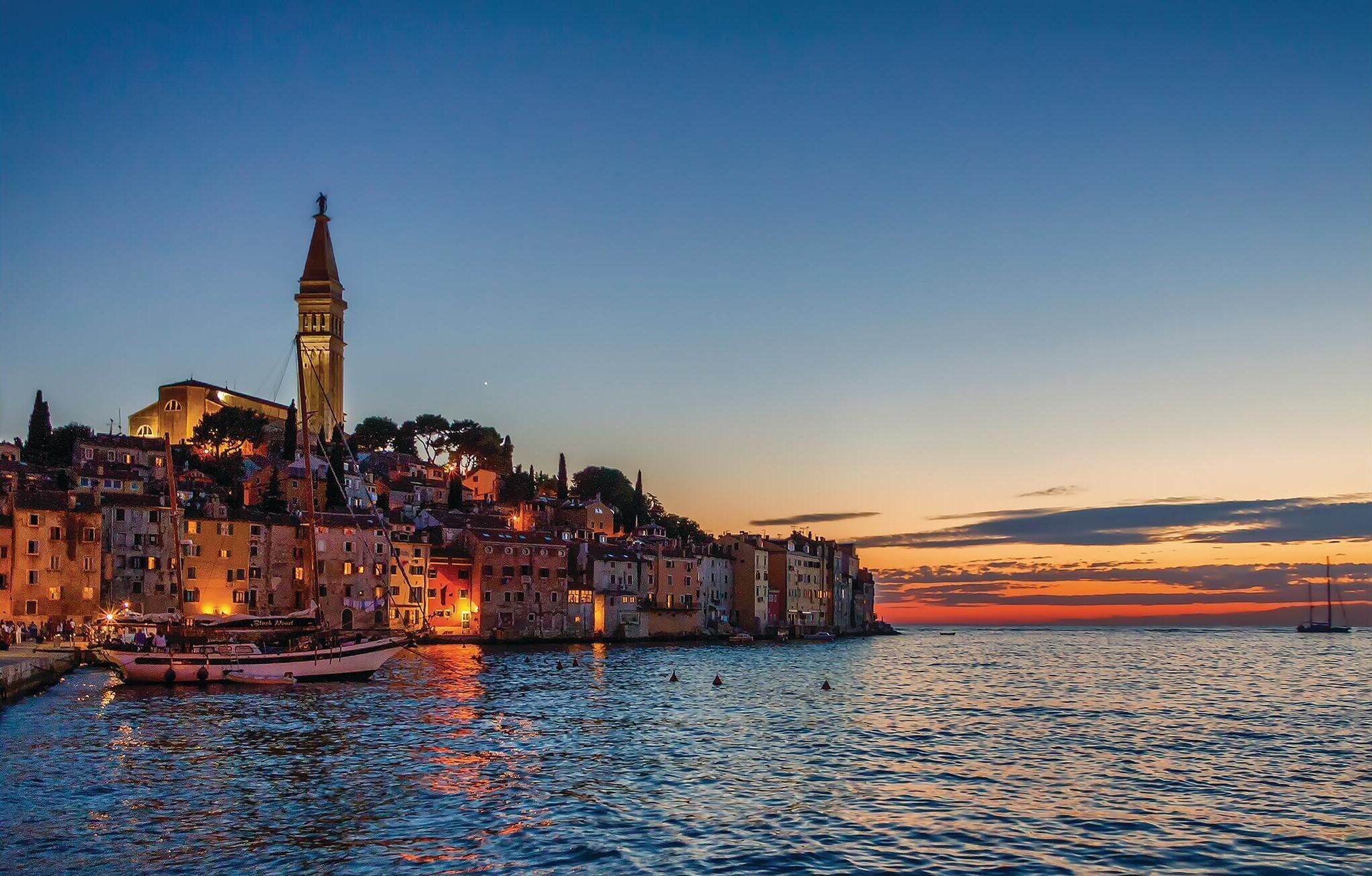 A coastal cityscape at dusk with a prominent tower, historic buildings, and a calm sea.