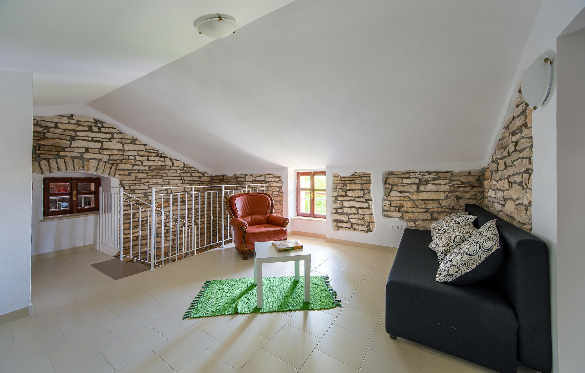 A living room with a stone wall, a black sofa, a red chair, a white table, and a green rug.
