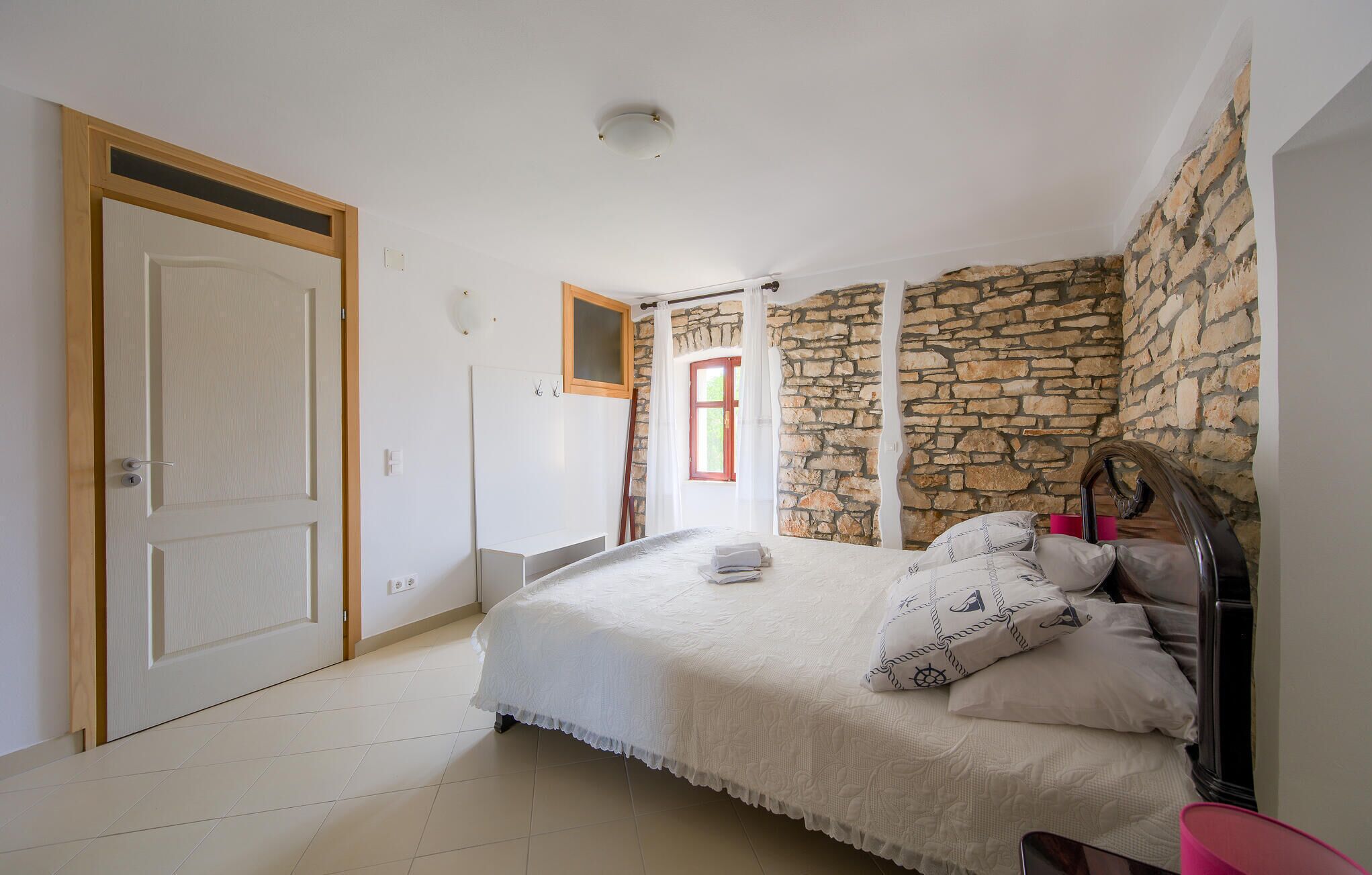 A bedroom with a stone wall, a bed with a white bedspread, a wooden door, and a small window with curtains.