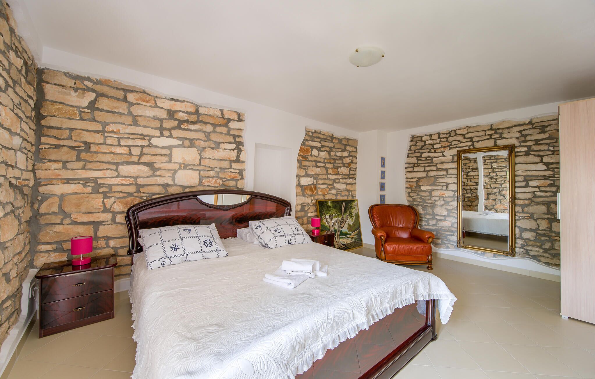 A bedroom with a stone wall, a wooden bed, a chair, and a mirror.