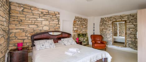 A bedroom with a stone wall, a wooden bed, a chair, and a mirror.