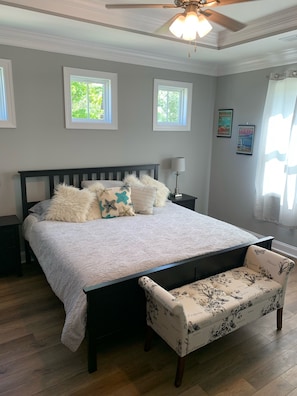 Quarto com cama, um banquinho, duas mesas de cabeceira, um ventilador de teto e duas janelas.