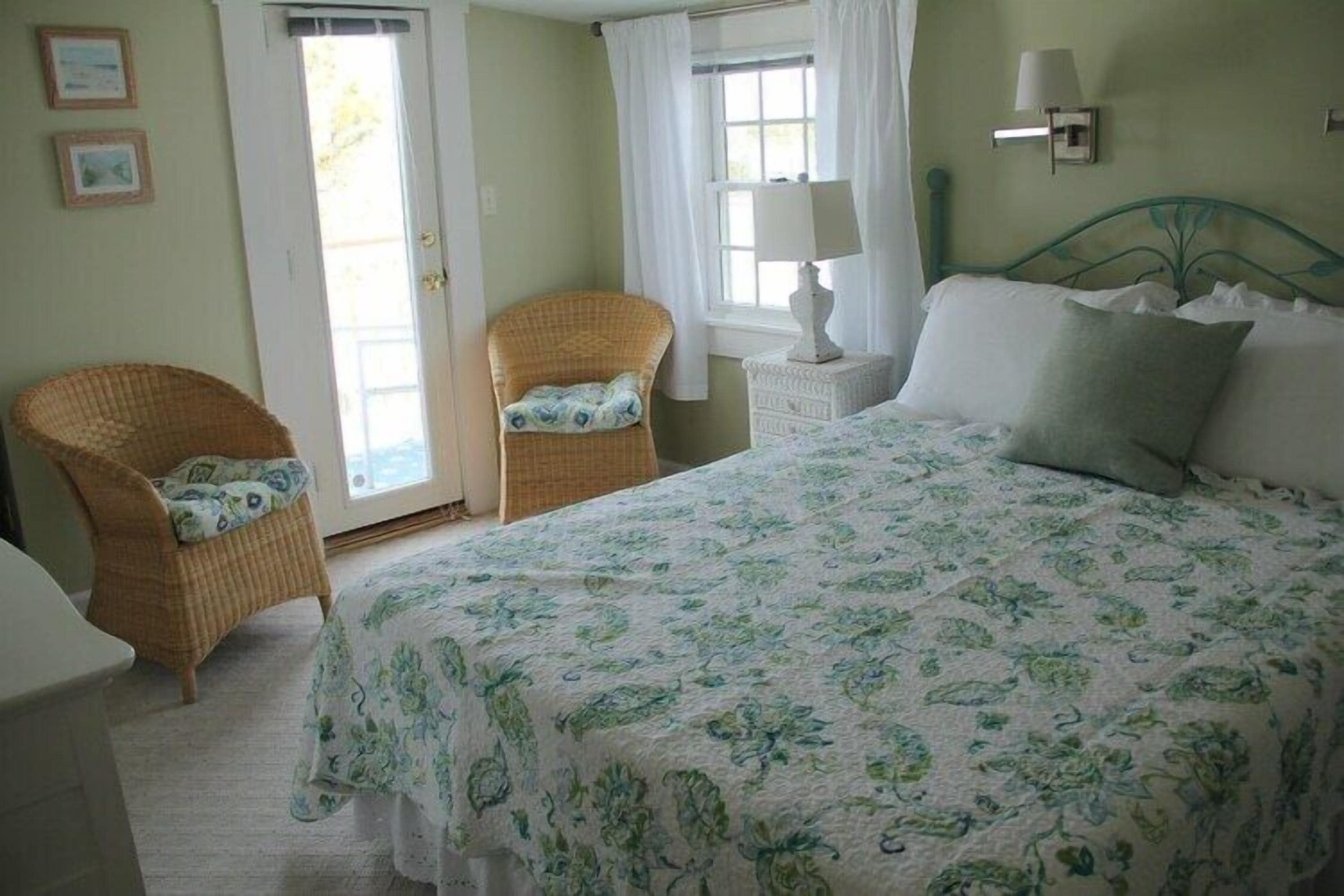 Un dormitorio con una cama, dos sillas de mimbre, una mesita de noche, una lámpara y una puerta que da a un balcón.