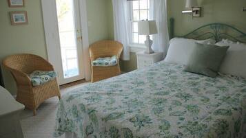 Un dormitorio con una cama, dos sillas de mimbre, una mesita de noche, una lámpara y una puerta que da a un balcón.