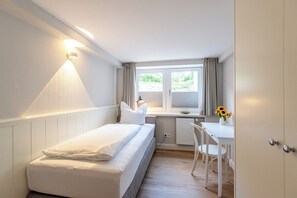 Ein kleiner, gut beleuchteter Raum mit einem Bett, einem Schreibtisch und einem Stuhl.