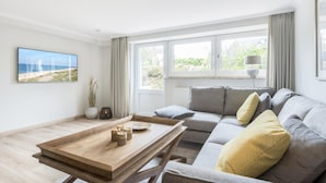 Ein modernes Wohnzimmer mit einem grauen Sofa, einem Holztisch und einem großen Fenster mit Vorhängen.