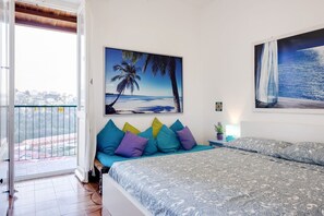Un dormitorio con cama, almohadas coloridas, un cuadro de playa y vistas de la ciudad desde un balcón.