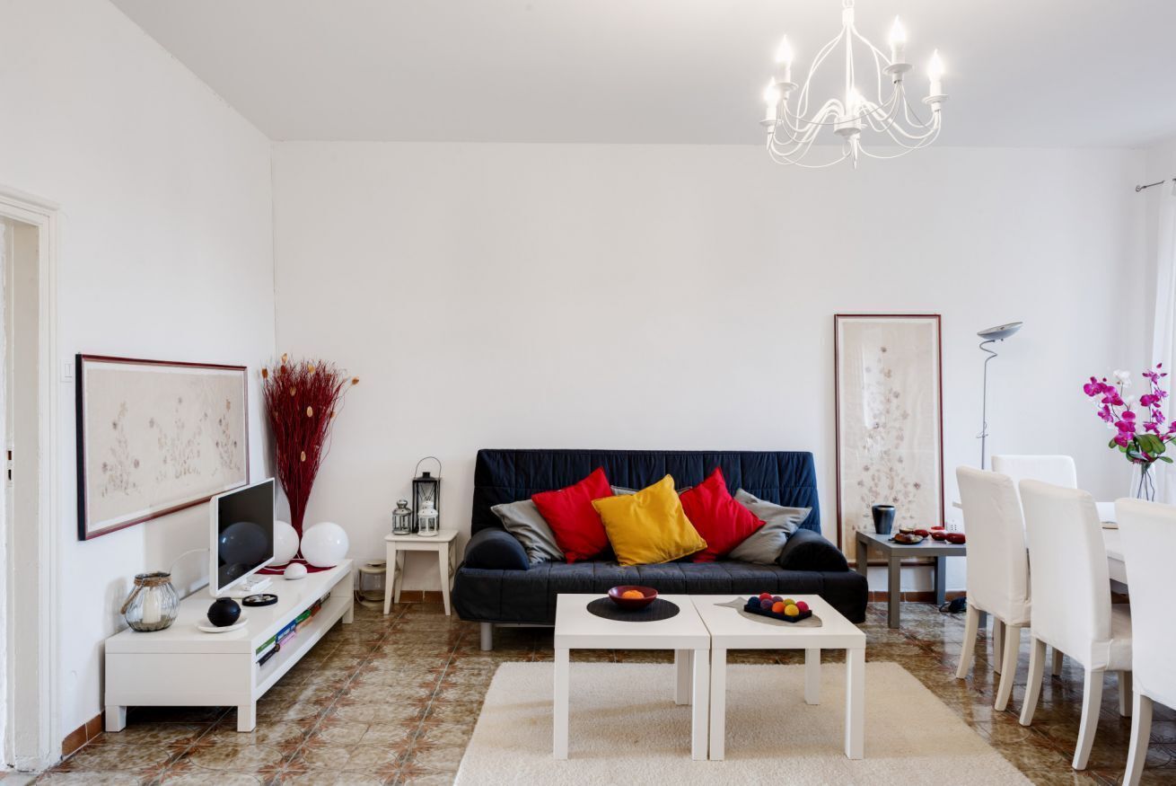 Una sala de estar con un sofá azul oscuro, mesas auxiliares blancas, una araña y un comedor con sillas blancas.