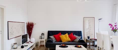 Una sala de estar con un sofá azul oscuro, mesas auxiliares blancas, una araña y un comedor con sillas blancas.