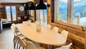 Un intérieur en bois avec une table ronde et des chaises blanches, de grandes fenêtres offrant une vue sur les montagnes et une horloge au mur.