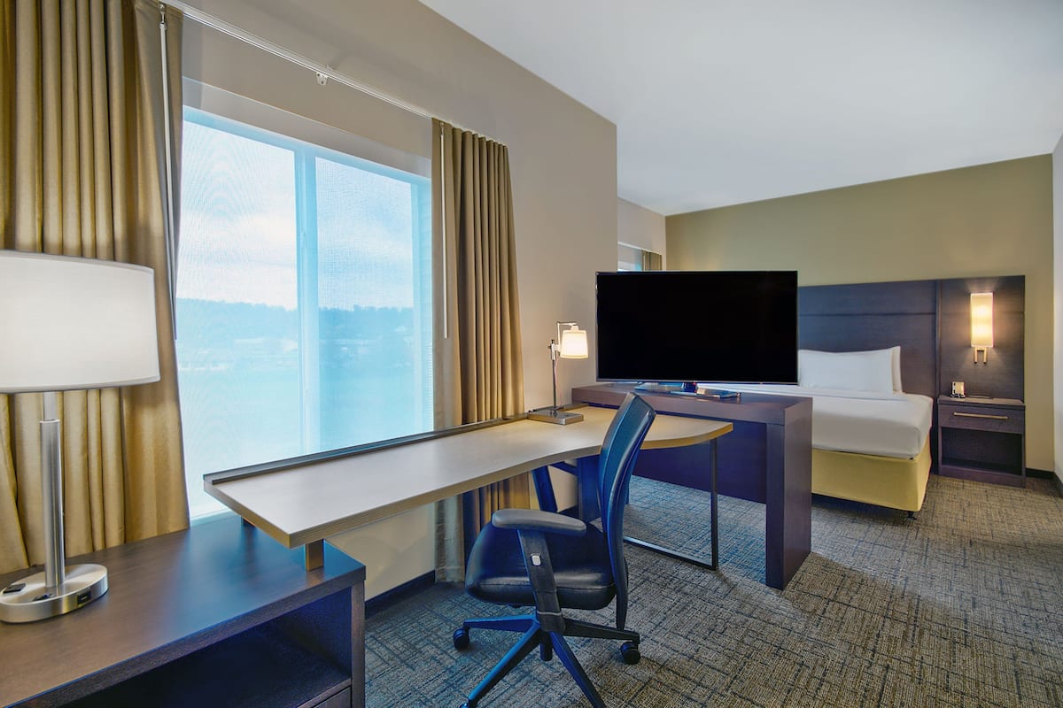 A hotel room with a desk, chair, bed, TV, and a view of the city.