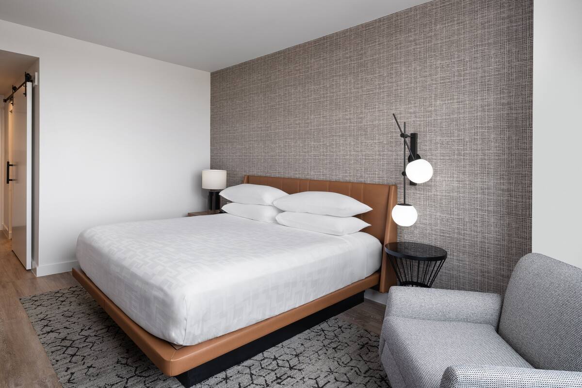 A modern hotel room with a large bed, a bedside table with a lamp, a small side table, and a sofa.