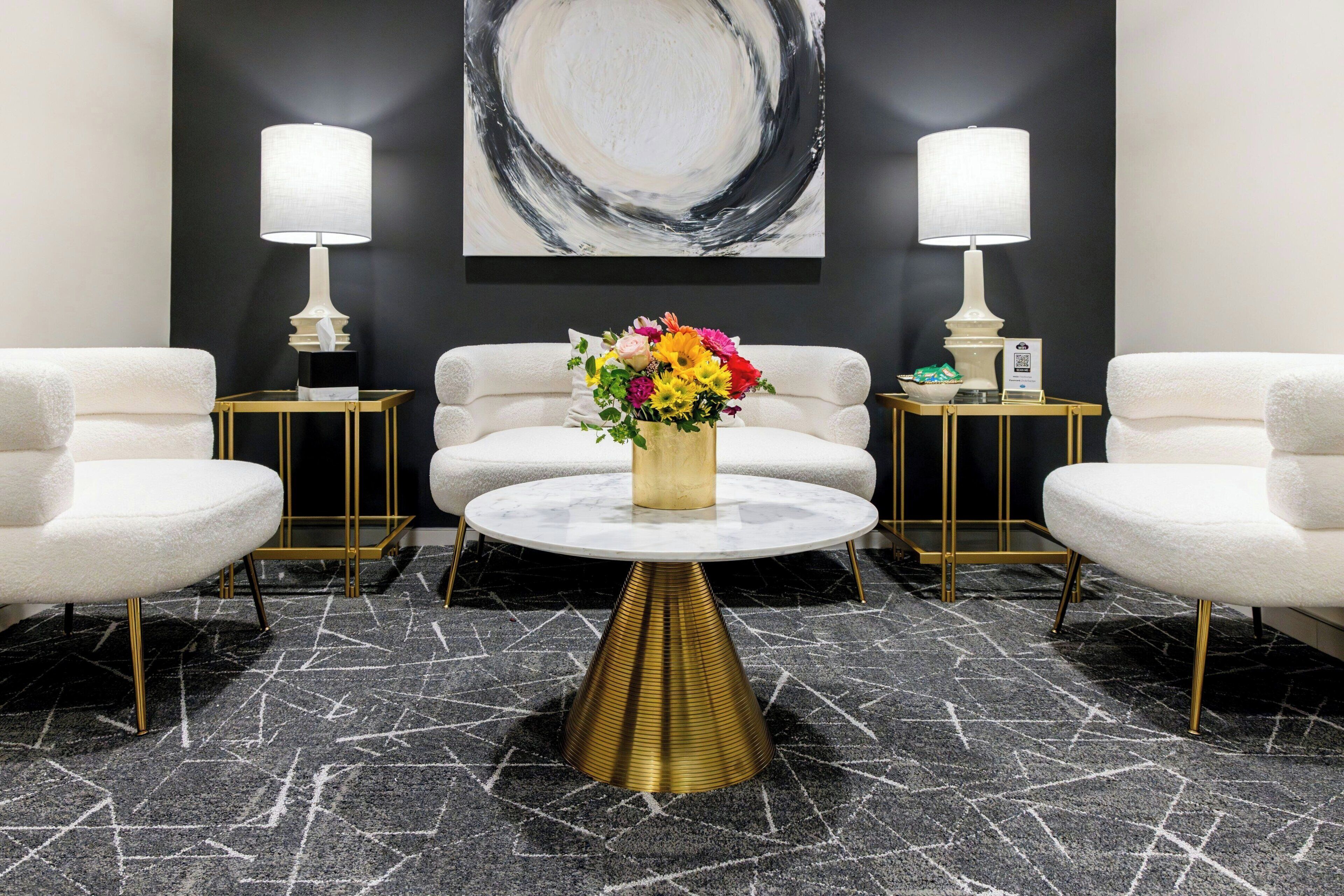 A modern lounge area with a round table, two white armchairs, and a gold accent table.
