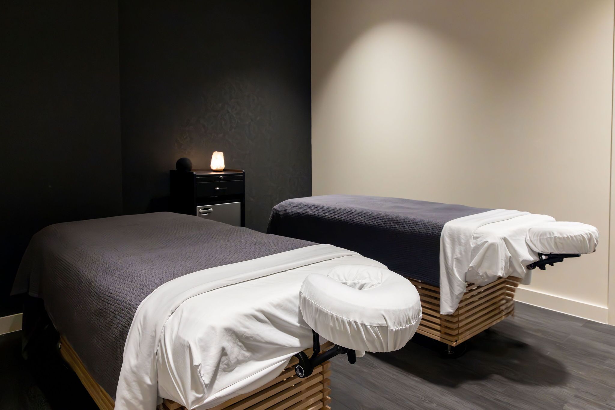Habitación de hotel con dos camas, cada una con un almohadón blanco y una colcha oscura. Hay una mesita de noche pequeña con una lámpara y un cajón.