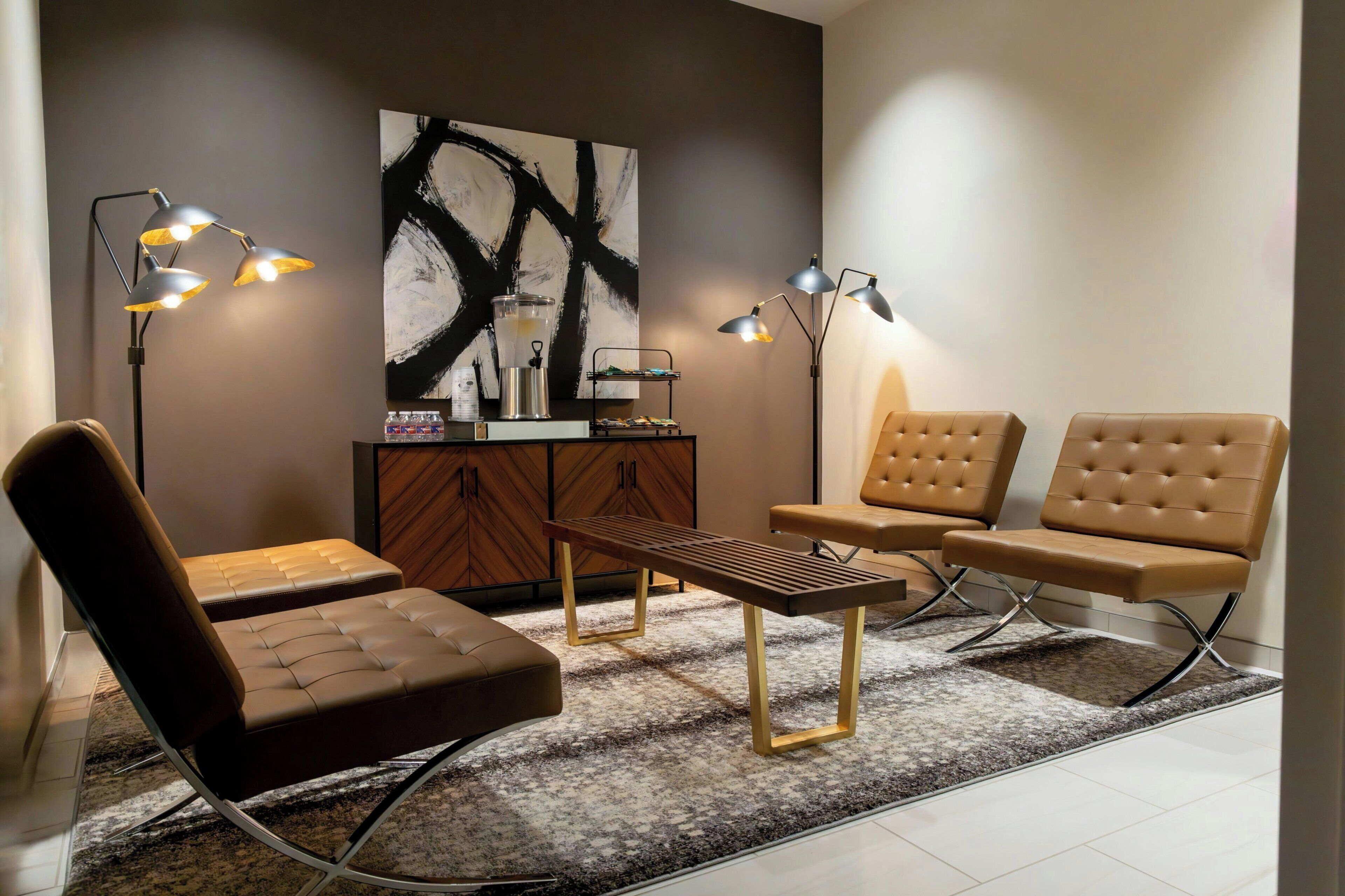 A modern living room with a brown leather sofa, two brown armchairs, a wooden coffee table, and two floor lamps. A large abstract painting and a wooden cabinet with a glass container are on the wall.