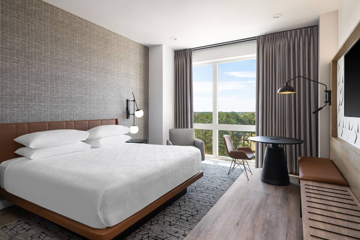 A modern hotel room with a large bed, a seating area with a table, and a view of greenery through the window.