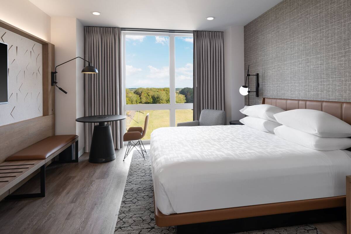 A modern hotel room with a large bed, a desk, a chair, and a view of the outdoors.