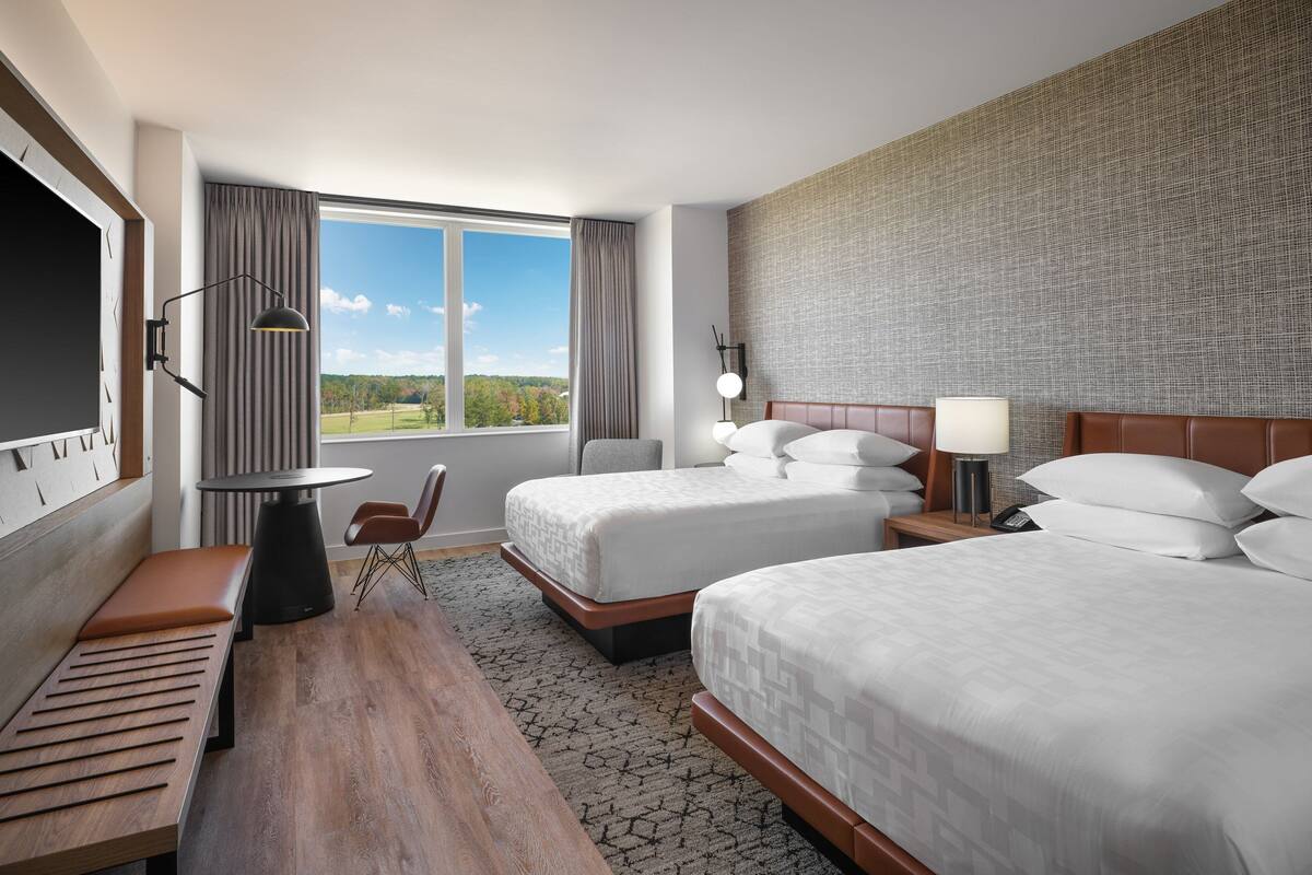 A hotel room with two beds, a desk, a chair, a TV, and a large window with a view of the outdoors.