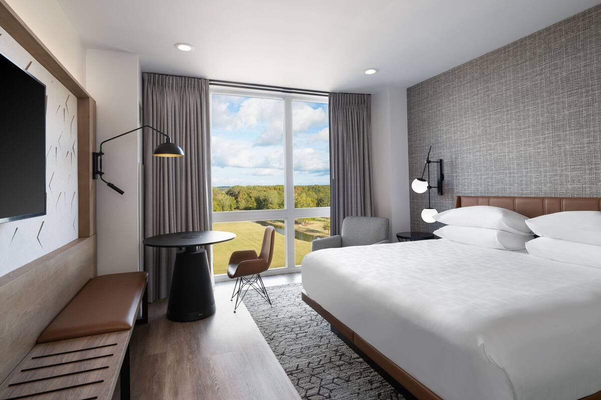 A modern hotel room with a large bed, a desk, a chair, and a view of the outdoors.