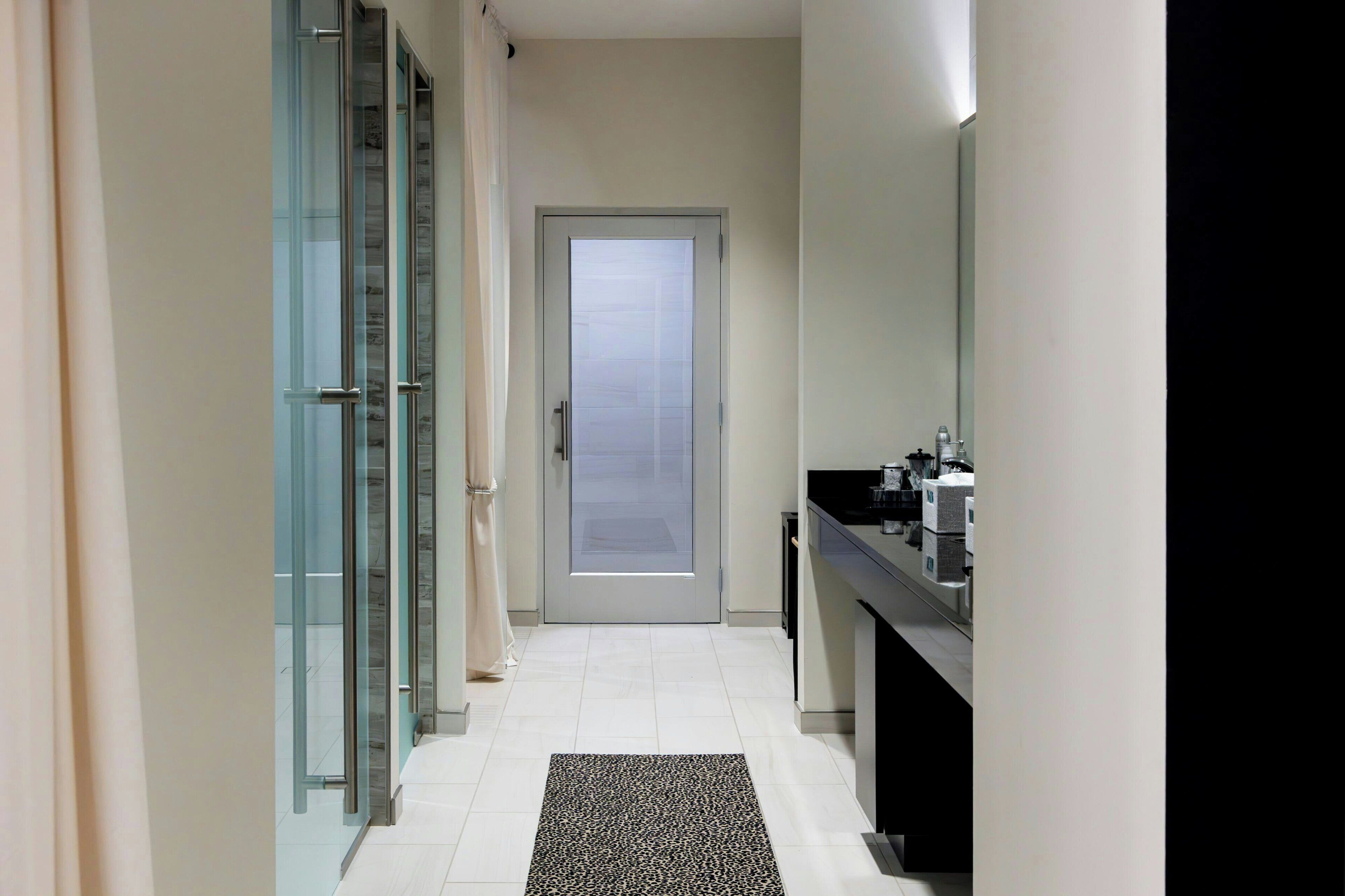 A narrow hallway with a glass door at the end, a black countertop with a mirror, and a patterned rug on the floor.