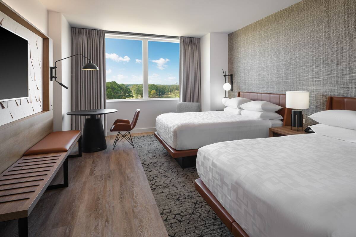 A hotel room with two beds, a desk, a chair, and a large window with a view of greenery.