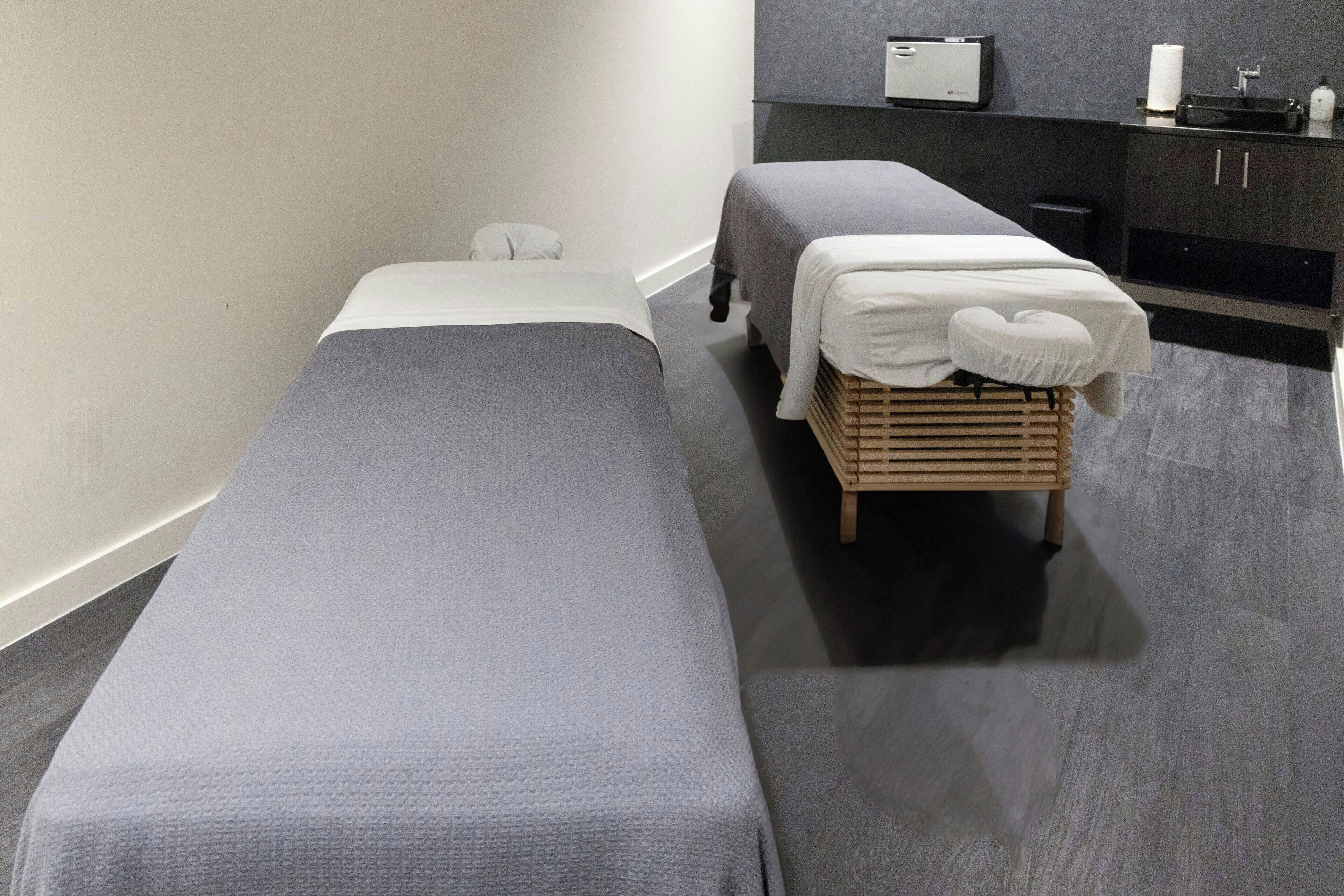 Two single beds with grey bedding, a wooden bench, a small white chair, a wall-mounted device, and a black cabinet with a sink.