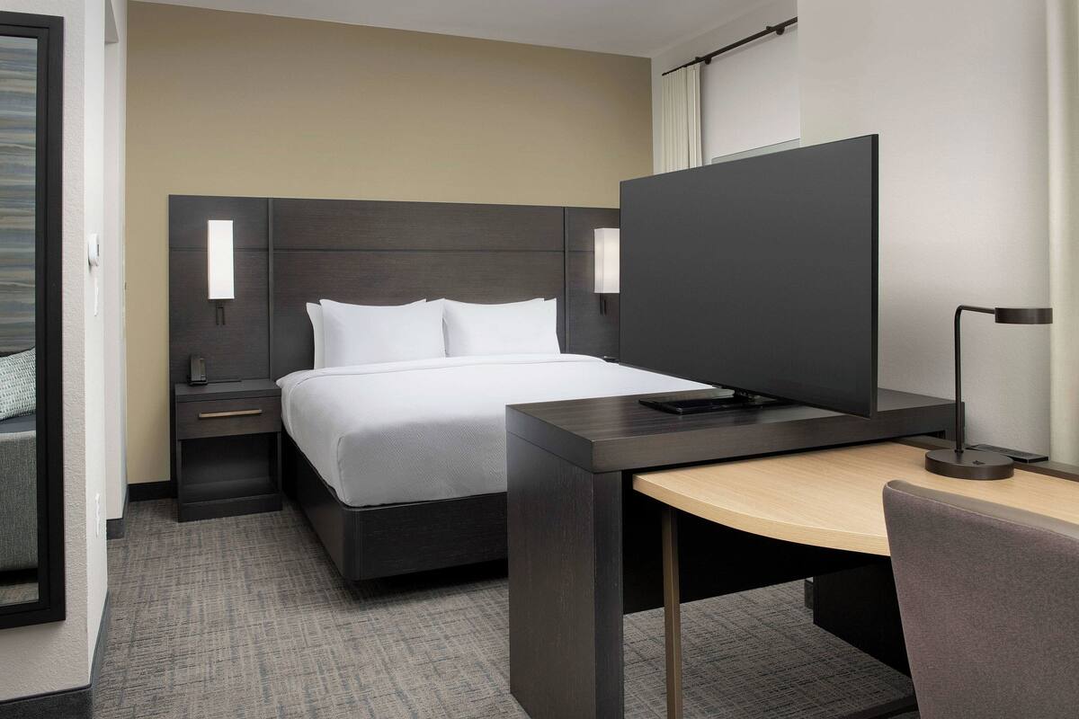 A modern hotel room with a bed, bedside table, desk, chair, and a television.