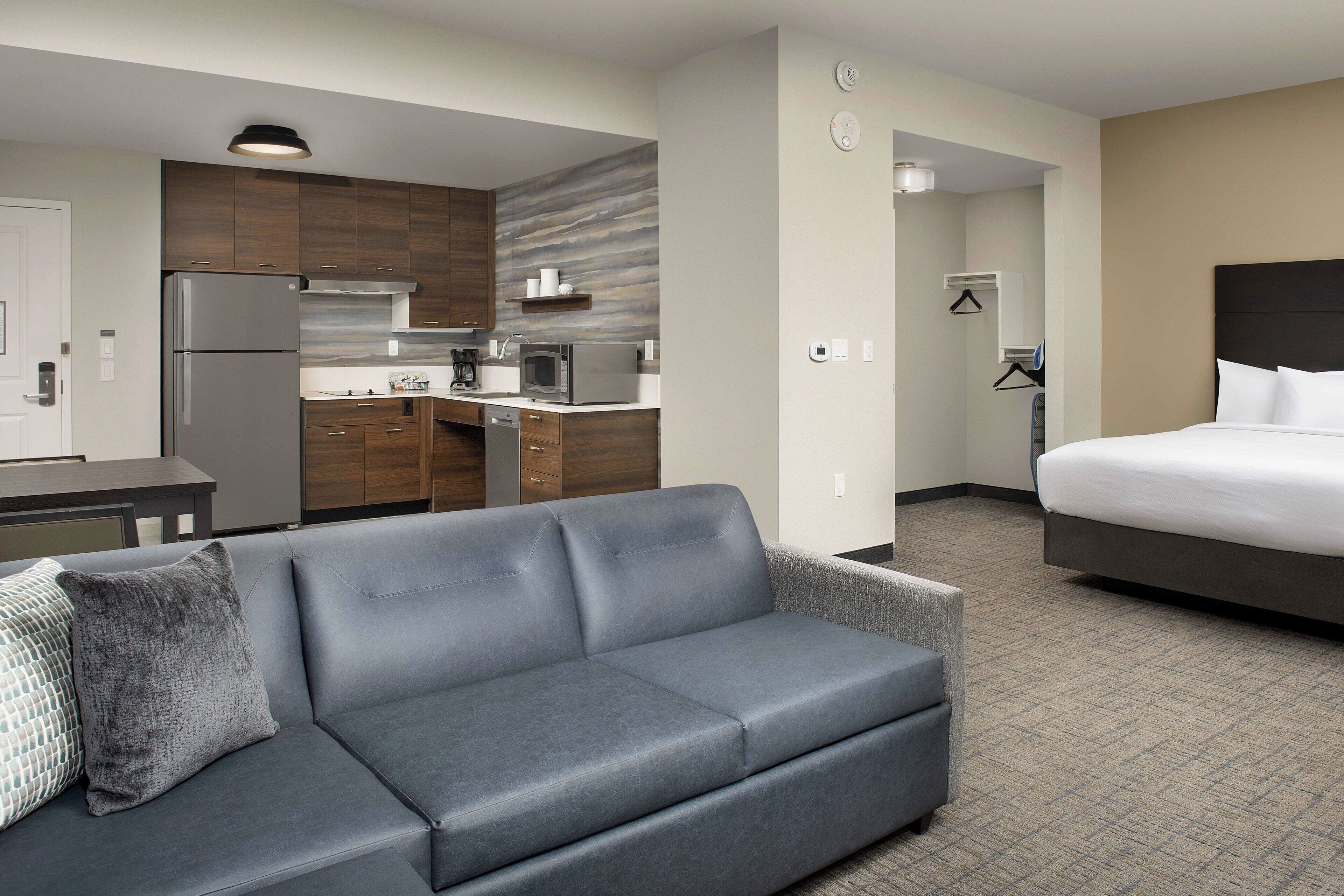 A modern hotel room with a grey sofa, a bed, a kitchenette, and a bathroom.
