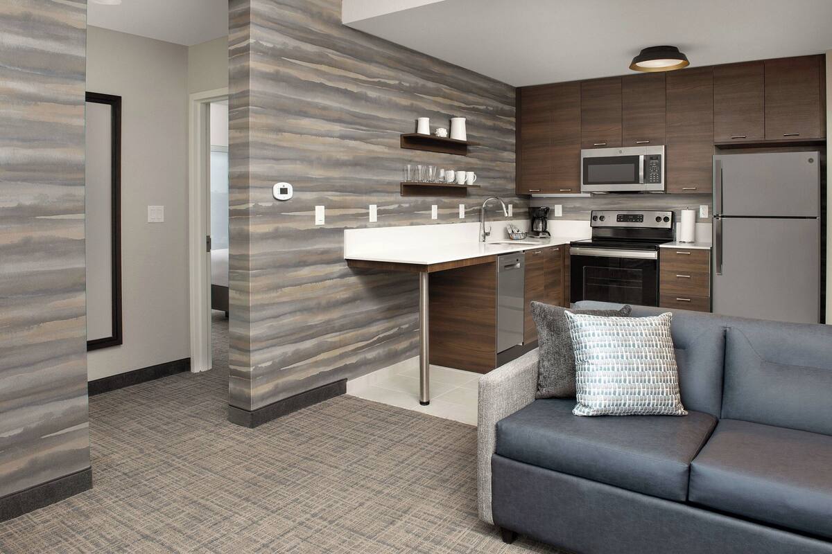 A modern kitchen with a built-in microwave, oven, and refrigerator. There is a gray sofa with a patterned pillow in the living area.