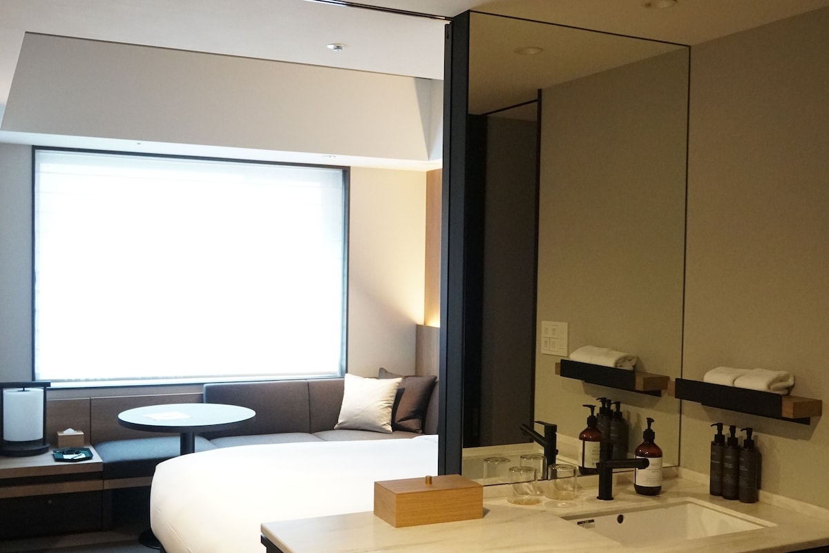A modern hotel room with a large bed, a round table, a built-in desk, a large mirror, and a bathroom with a vanity and shelves.