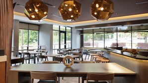 A modern dining area with wooden tables and chairs, large windows, and geometric pendant lighting. - SpringHill Suites by Marriott Durham City View (Durham)