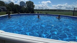 An outdoor swimming pool with clear blue water, surrounded by a grassy area and trees. - Country living with a pool, fire pit & family-friendly! (Fond du Lac)
