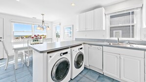 A modern kitchen with white cabinets, a built-in washer and dryer, a large window with a view, and a dining area with white chairs. - Beach Front! (Long Beach)