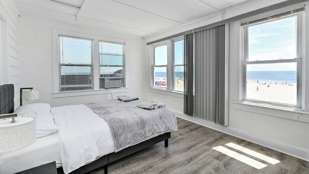 A bedroom with a bed, a bedside table, a lamp, a window with a view of the beach, and a ceiling fan. - Modern Beachfront Suite 202 (Ocean City)