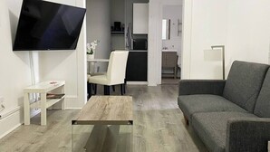 A modern living room with a flat-screen TV, a grey sofa, a coffee table, and a dining area with a chair and a small table. - Modern Beachfront Suite 202 (Ocean City)