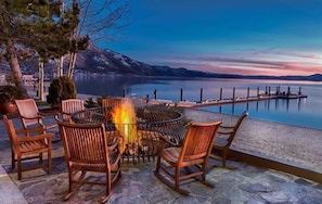 A terrace with a circular table and chairs, overlooking a calm lake and distant mountains.