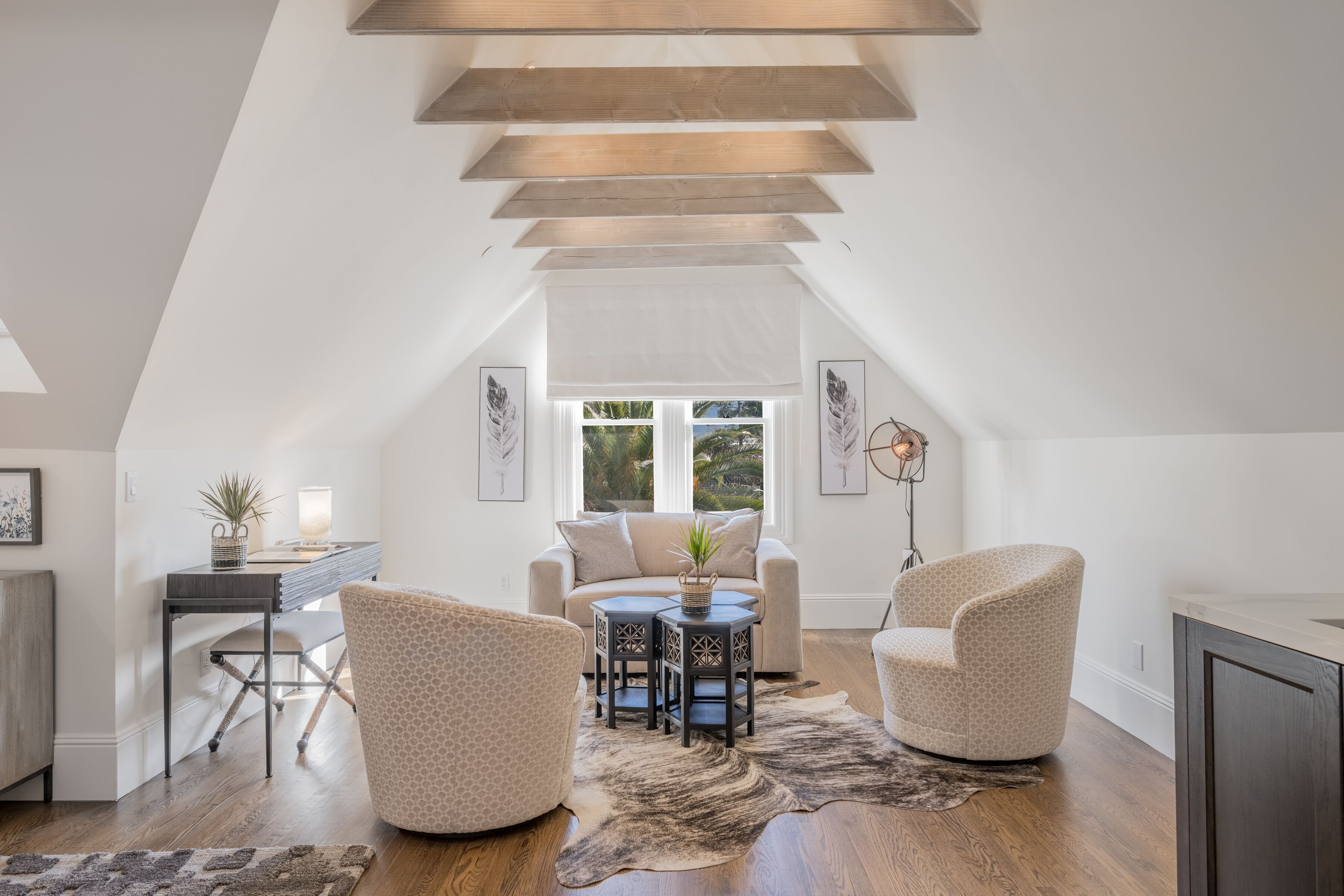 Ruime loft slaapkamer met een schuine kap, een groot raam, een gezellige zithoek met een bank en stoelen, en een kleine eettafel.