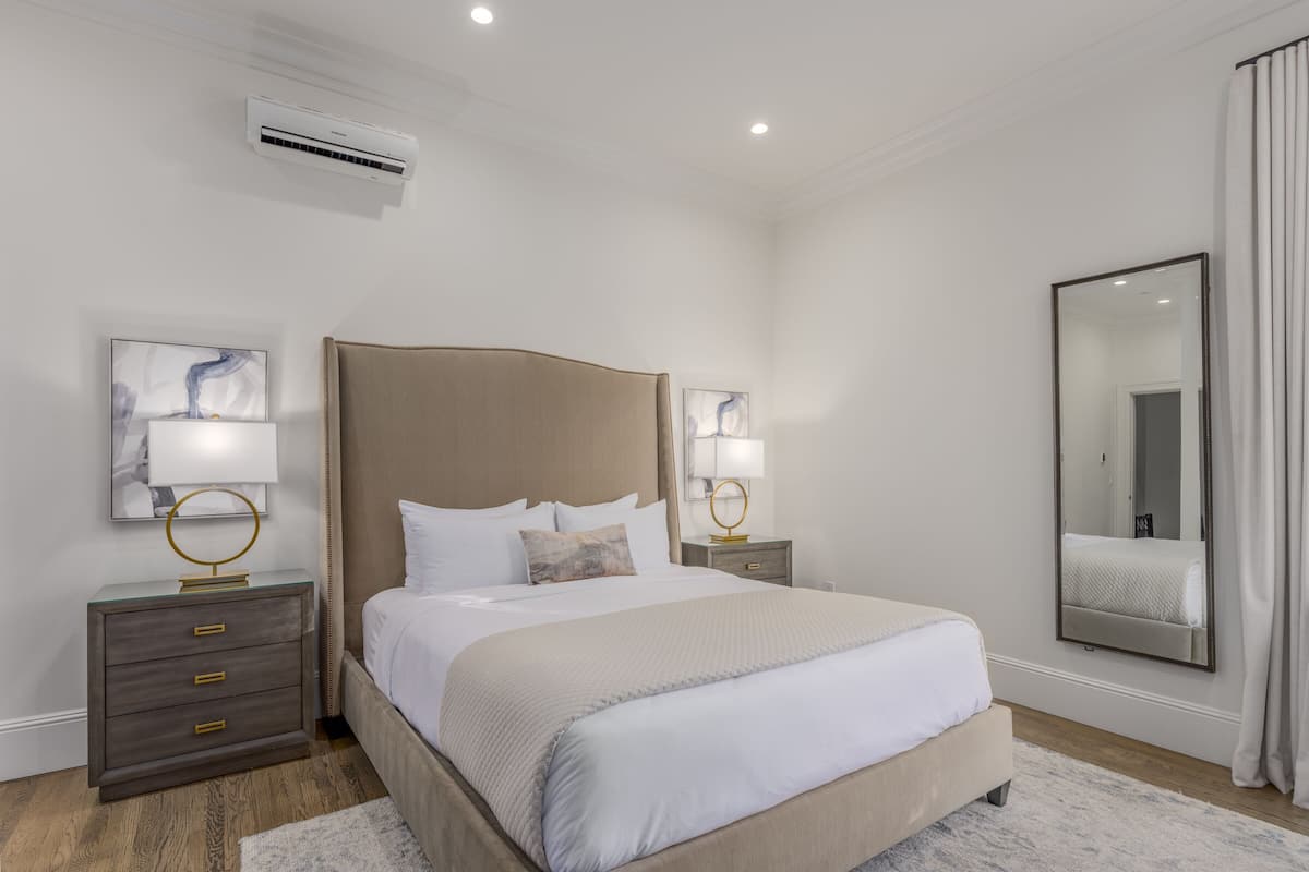 A modern bedroom with a large bed, two bedside tables with lamps, a wall-mounted air conditioner, and a large mirror.