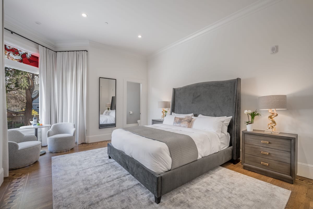 A modern bedroom with a large bed, a bedside table with lamps, a chair, and a view of the outdoors through a window with curtains.