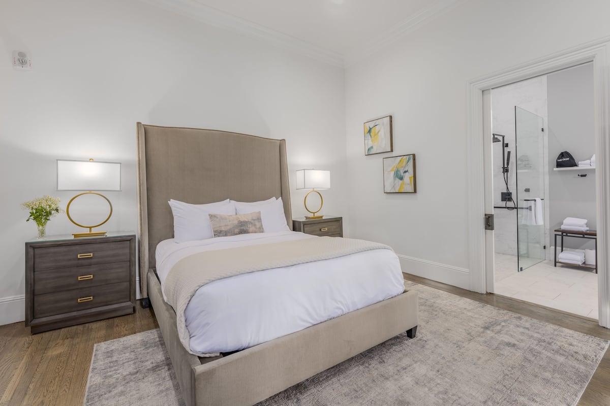 A modern bedroom with a large bed, bedside tables with lamps, a nightstand with a vase, and a glass shower door.