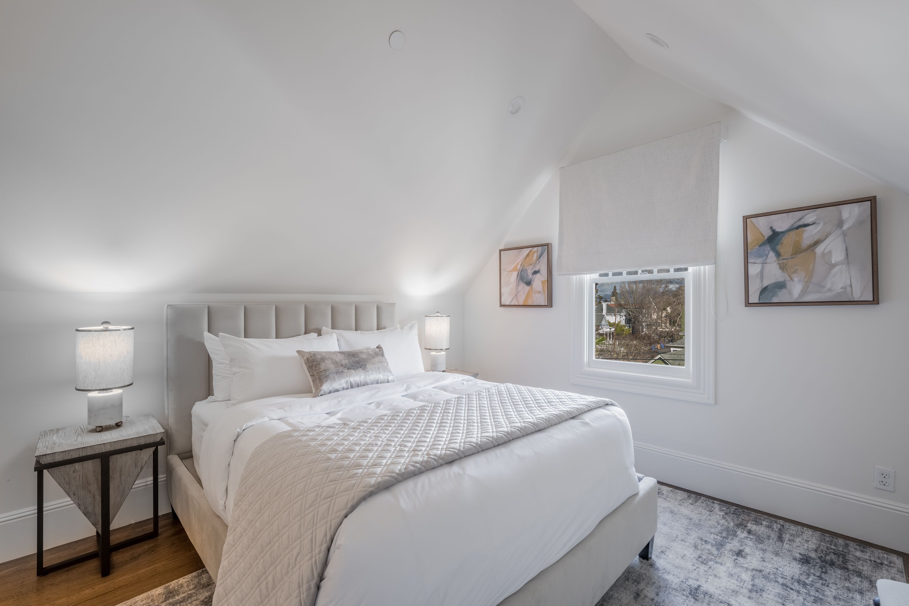 A bedroom with a large bed, a nightstand, a window, and two wall-mounted artworks.