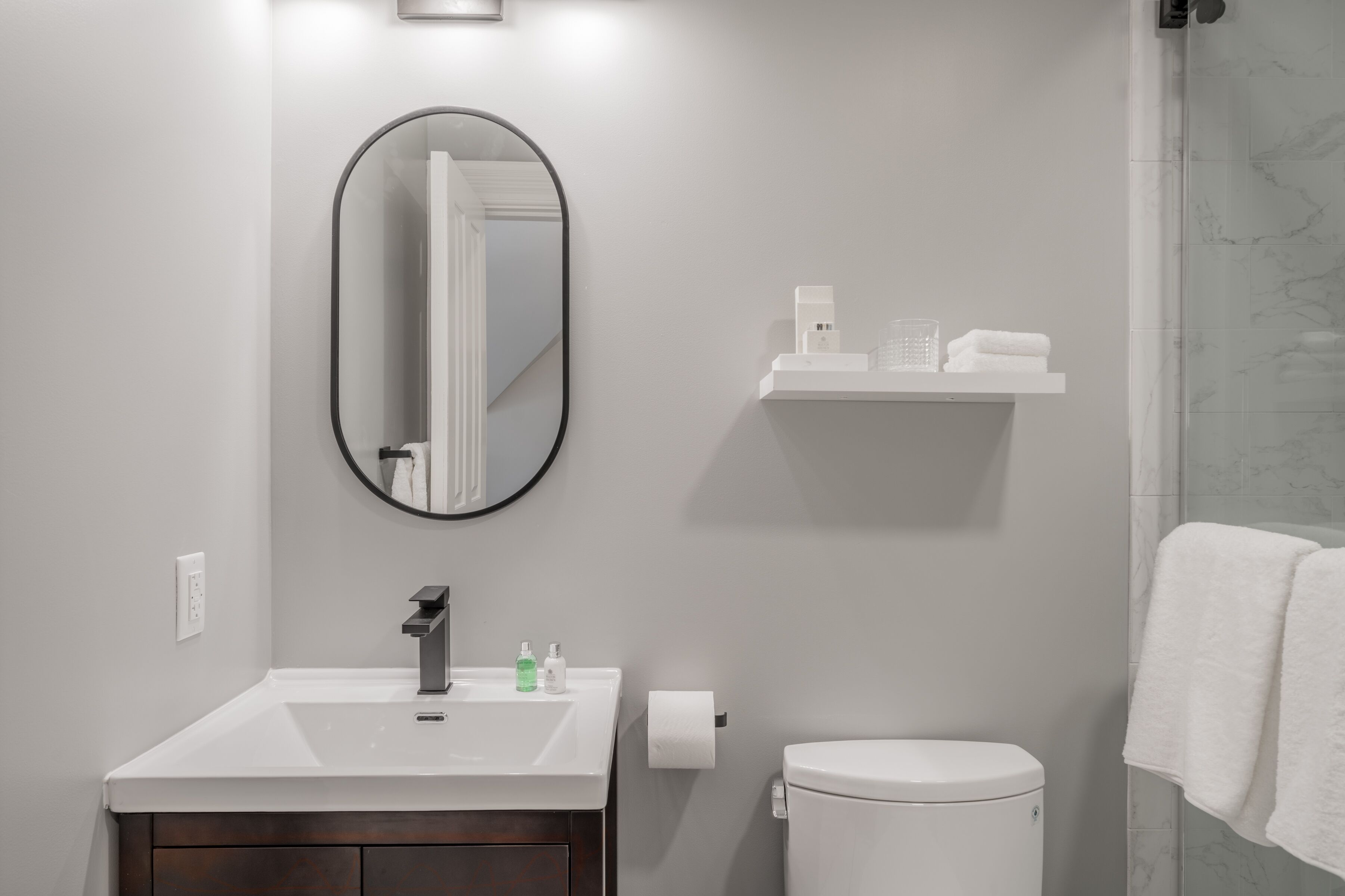 A bathroom with a white sink, mirror, and toilet. There is a shelf above the sink with toiletries.